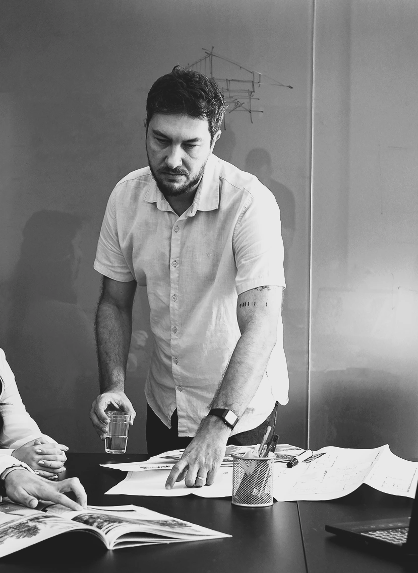 Adriano Couto no escritório, apresentando desenhos de arquitetura sobre a mesa, em fotografia preto e branco