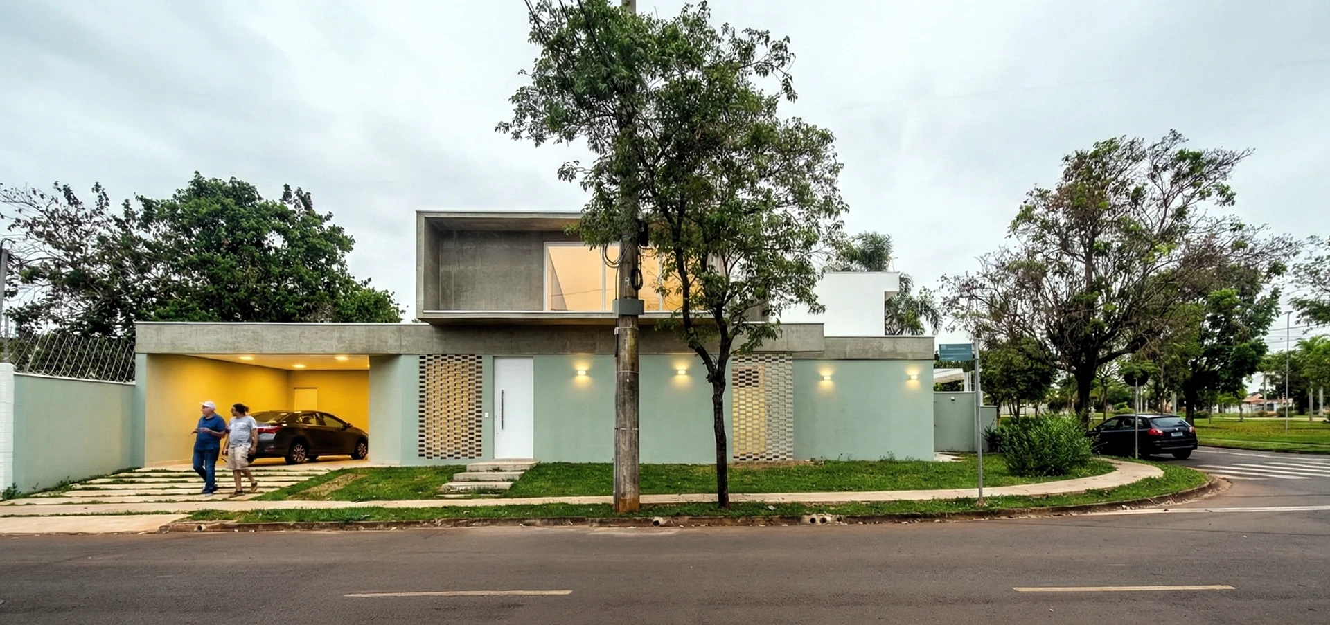 Casa Pátio: vista frontal da fachada iluminada na esquina, com jardim e pessoas na calçada