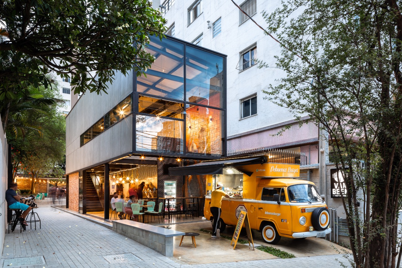 Cervejaria Van Been ao entardecer: fachada envidraçada iluminada, árvores e convivência na rua